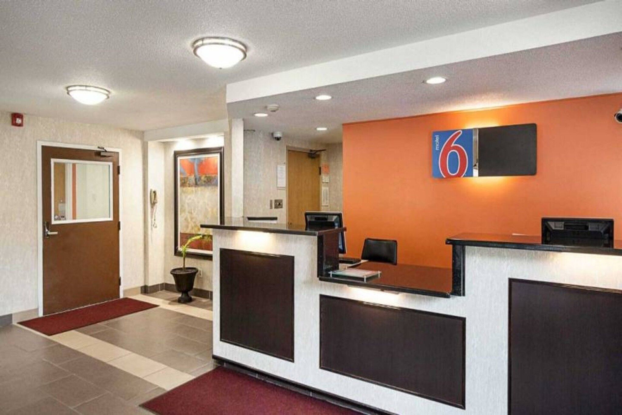 A hotel reception area with a reception desk, a number sign, a potted plant, and a red carpet.