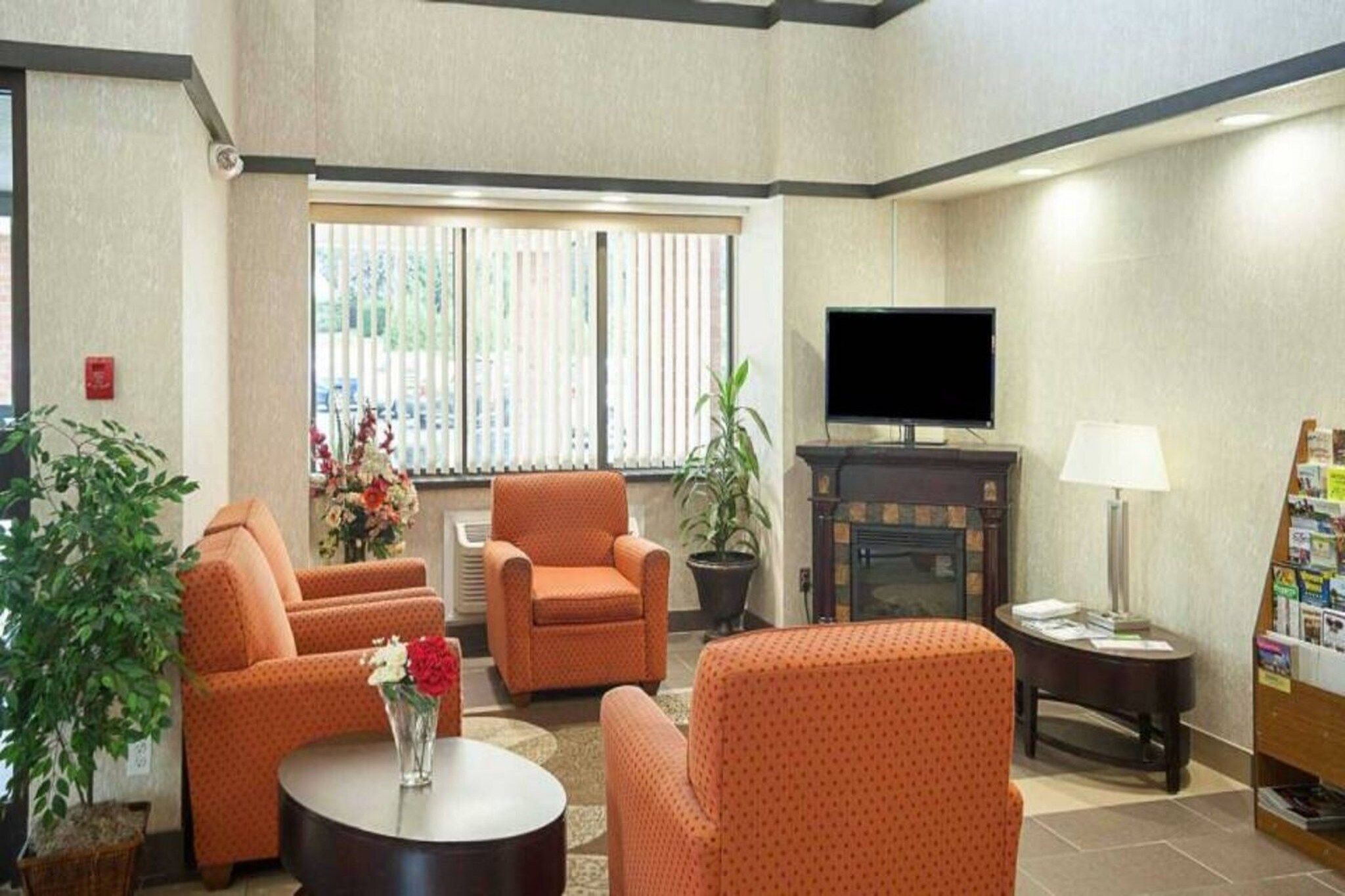 Um lounge de hotel com sofás de padrão laranja, uma TV de tela plana, uma lareira e uma pequena mesa com um vaso de flores.