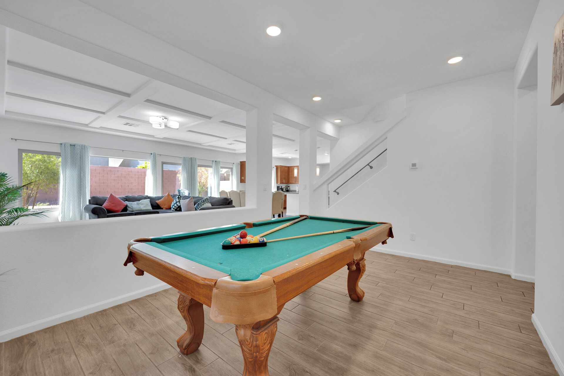 A spacious indoor area with a pool table, wooden flooring, and a view of a brick wall and greenery outside.