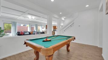 A spacious indoor area with a pool table, wooden flooring, and a view of a brick wall and greenery outside.