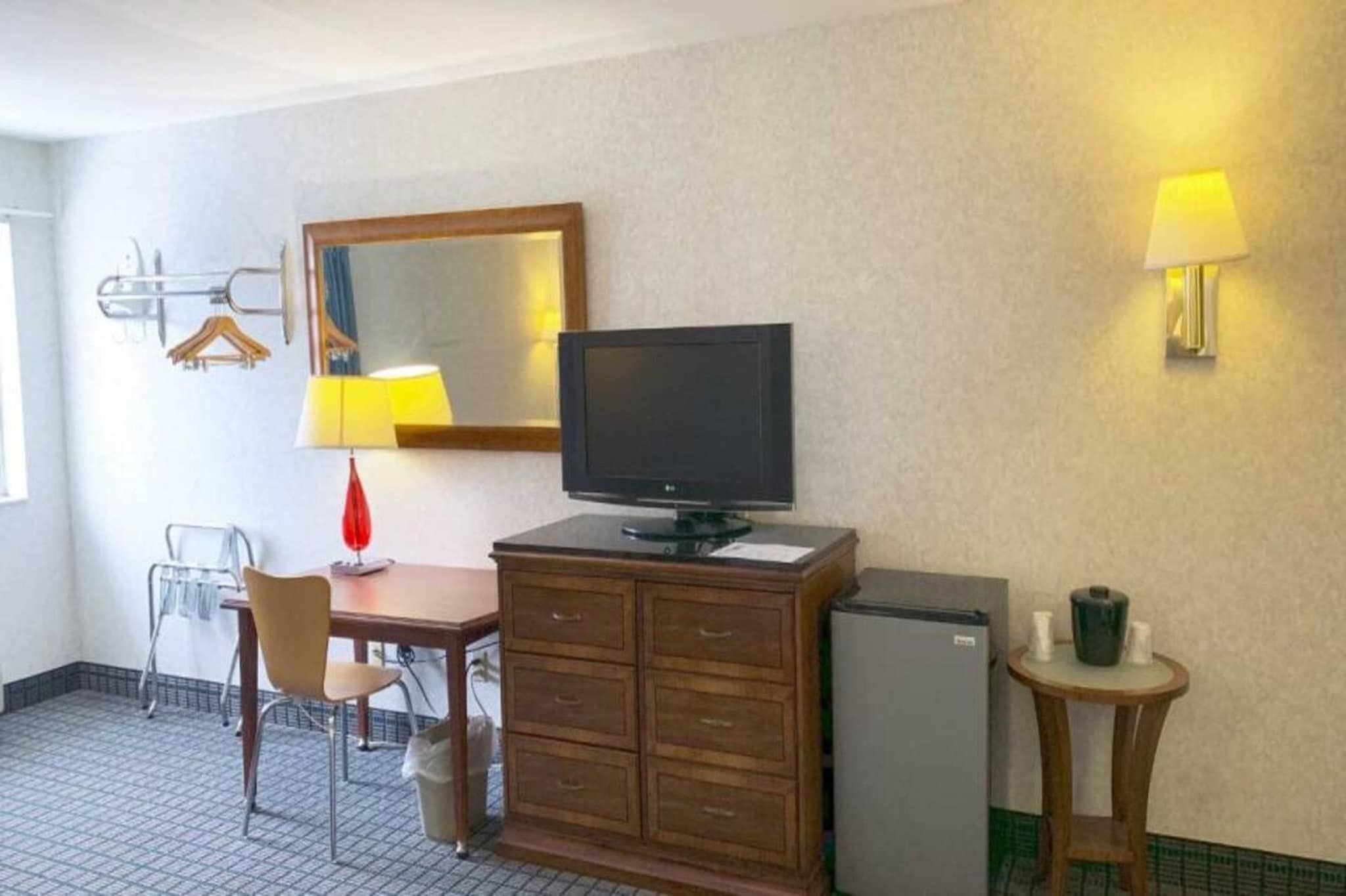 Habitación de hotel con escritorio, televisión, refrigerador, silla, lámpara y espejo.