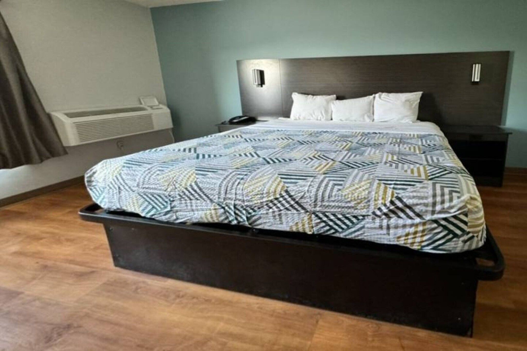 A hotel room with a bed, a bedside table, a nightstand, and a wall-mounted air conditioning unit.