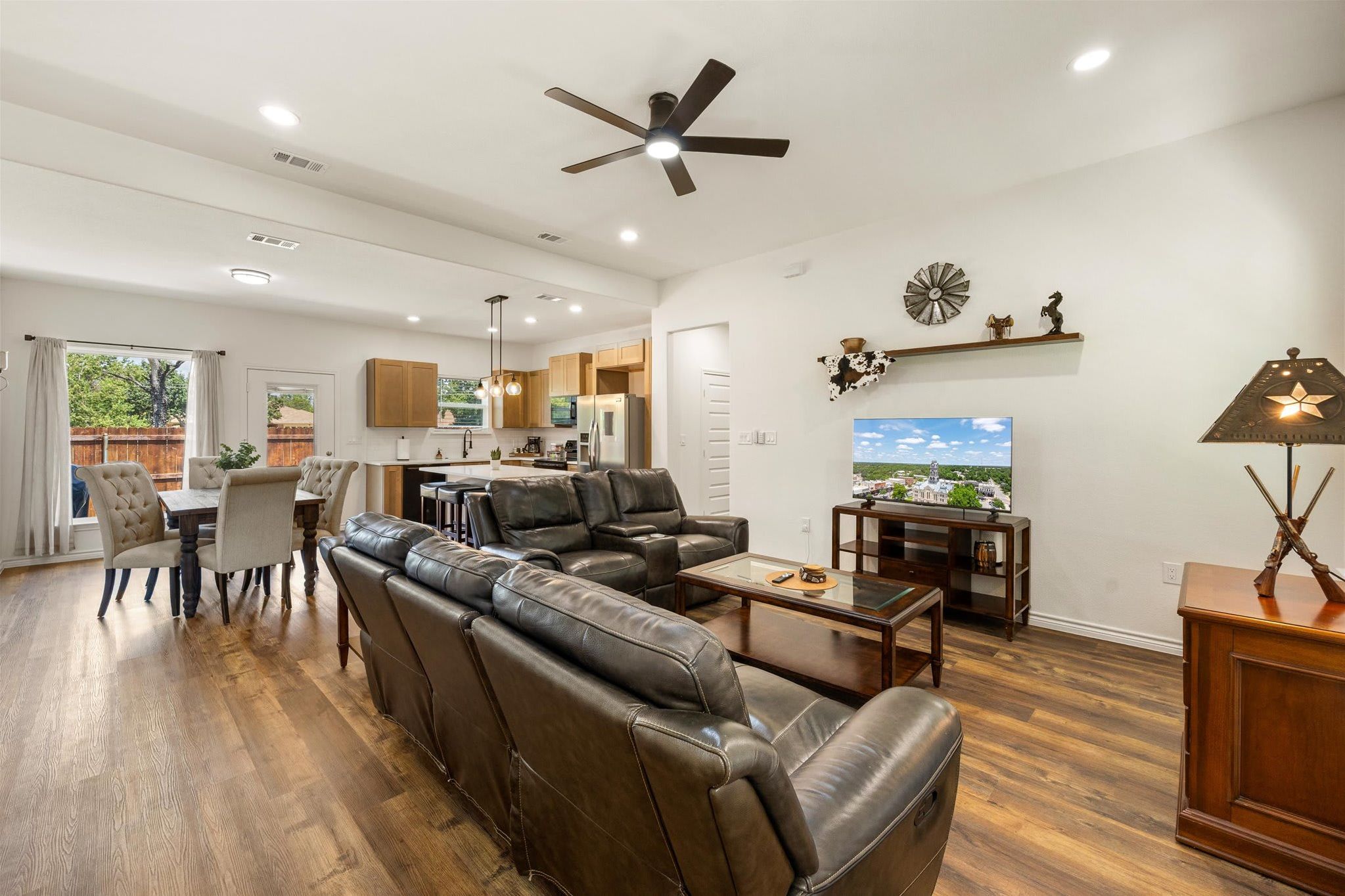 A spacious living room with a leather sofa, a dining area with a table and chairs, a kitchen with wooden cabinets, and a ceiling fan.