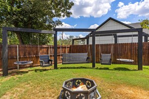 Een achtertuin met een houten pergola, twee schommels, een vuurkorf en een houten omheining.