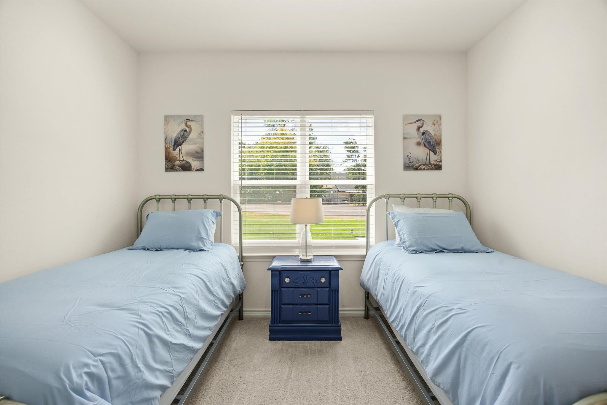 A bedroom with two twin beds, a bedside table with a lamp, and a window with blinds.