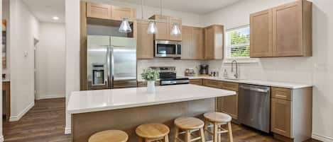 A modern kitchen with a central island, stainless steel appliances, wooden bar stools, and pendant lighting.