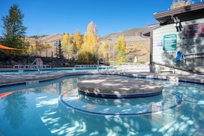 A swimming pool with a circular hot tub, surrounded by a fence and trees. There is a building with a sign and a bike rack.