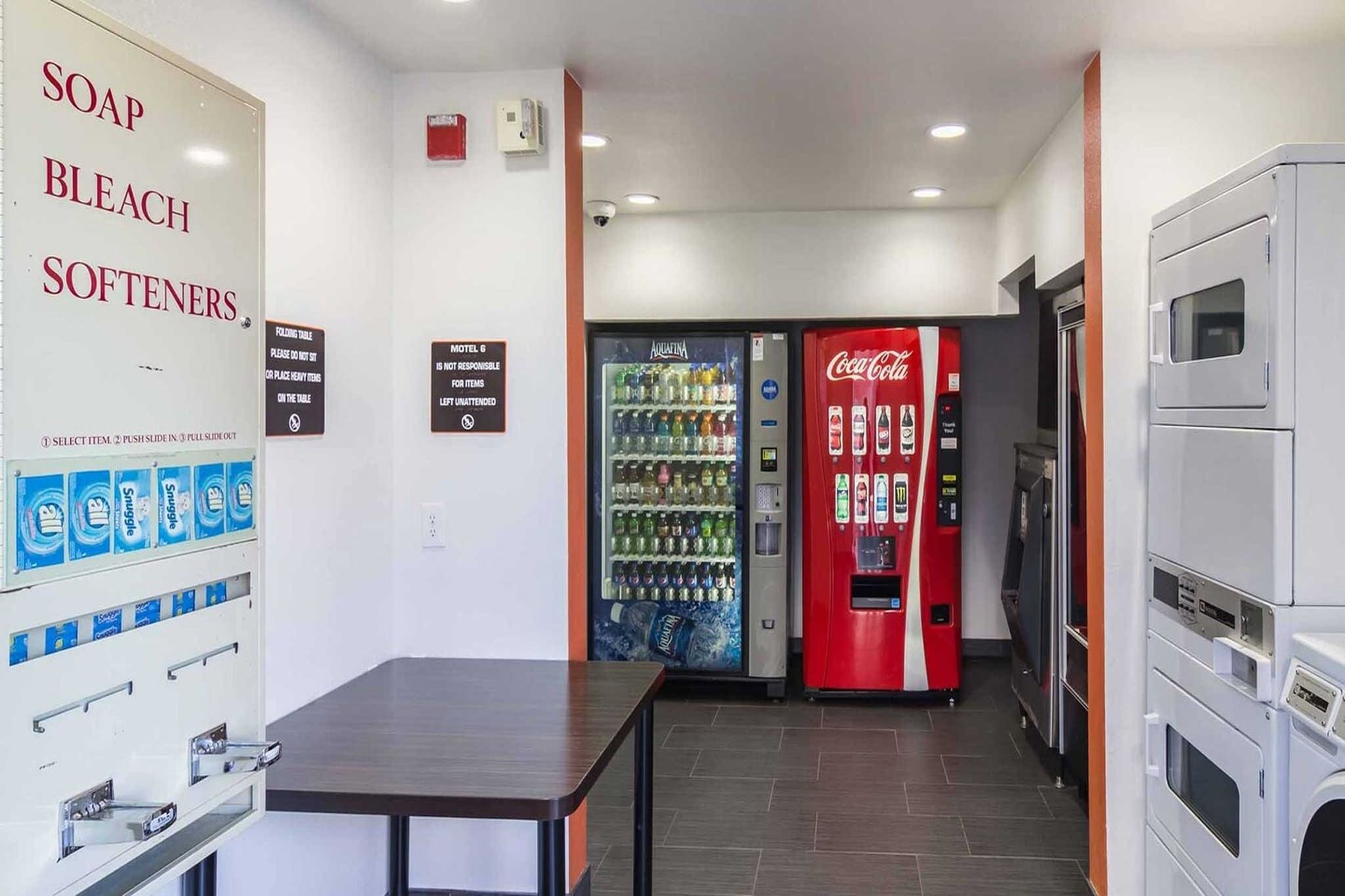 Una habitación de hotel con una mesa, una máquina expendedora de bebidas, una máquina de Coca-Cola y un dispensador de jabón y lejía instalado en la pared.