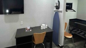 Een kleine kamer met een bureau, twee stoelen, een flatscreen televisie aan de muur, een haardroger en een klein strijkplank.