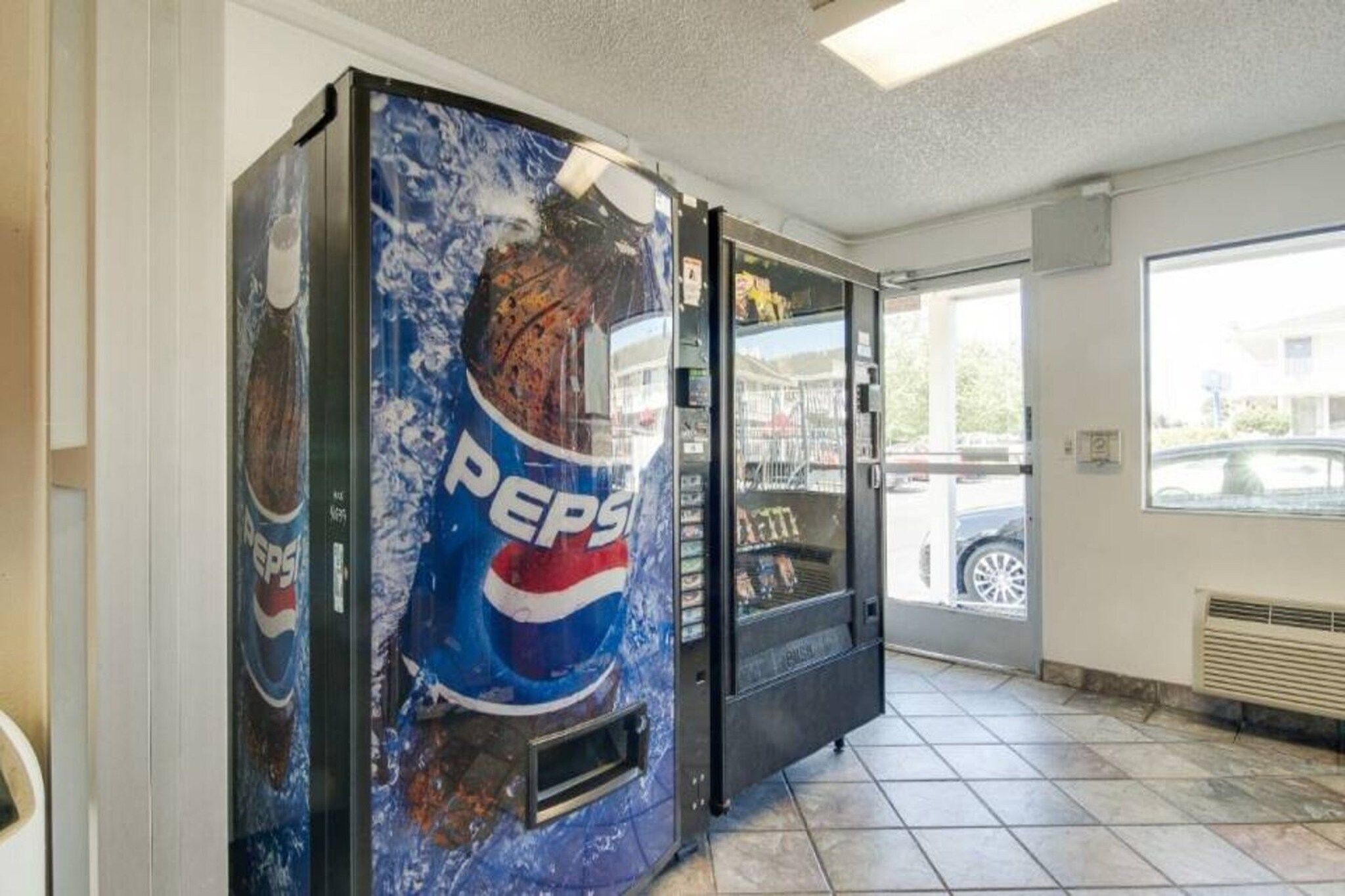 Un distributeur automatique orné du logo Pepsi, situé à l’intérieur d’un bâtiment, avec un sol carrelé et une fenêtre donnant sur l’extérieur, où l’on peut voir une voiture.