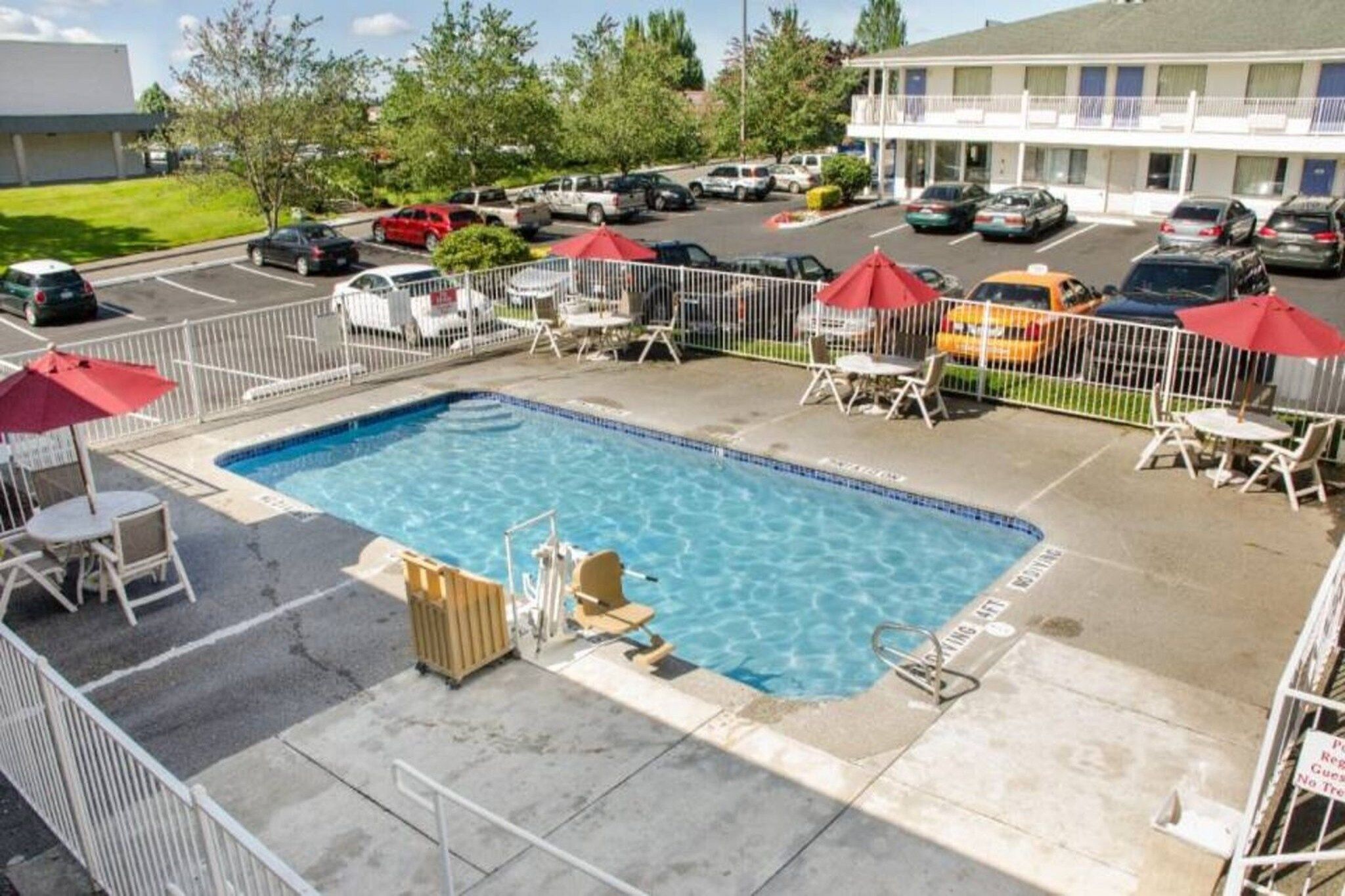 Une piscine dotée de chaises longues et de parasols, entourée d’une zone délimitée par une clôture, avec un parking en arrière-plan.