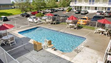 Une piscine dotée de chaises longues et de parasols, entourée d’une zone délimitée par une clôture, avec un parking en arrière-plan.