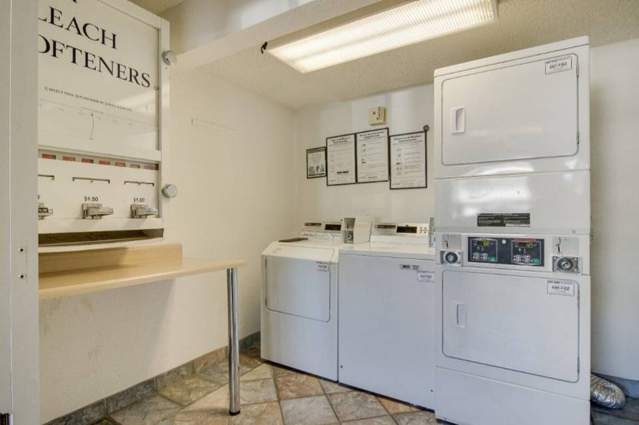 Une buanderie équipée d’une pile de lave-linge et de sèche-linge, d’un étendoir en métal et d’une affiche faisant la publicité de « LEACH OFTENERS » au prix de 1,50 $.