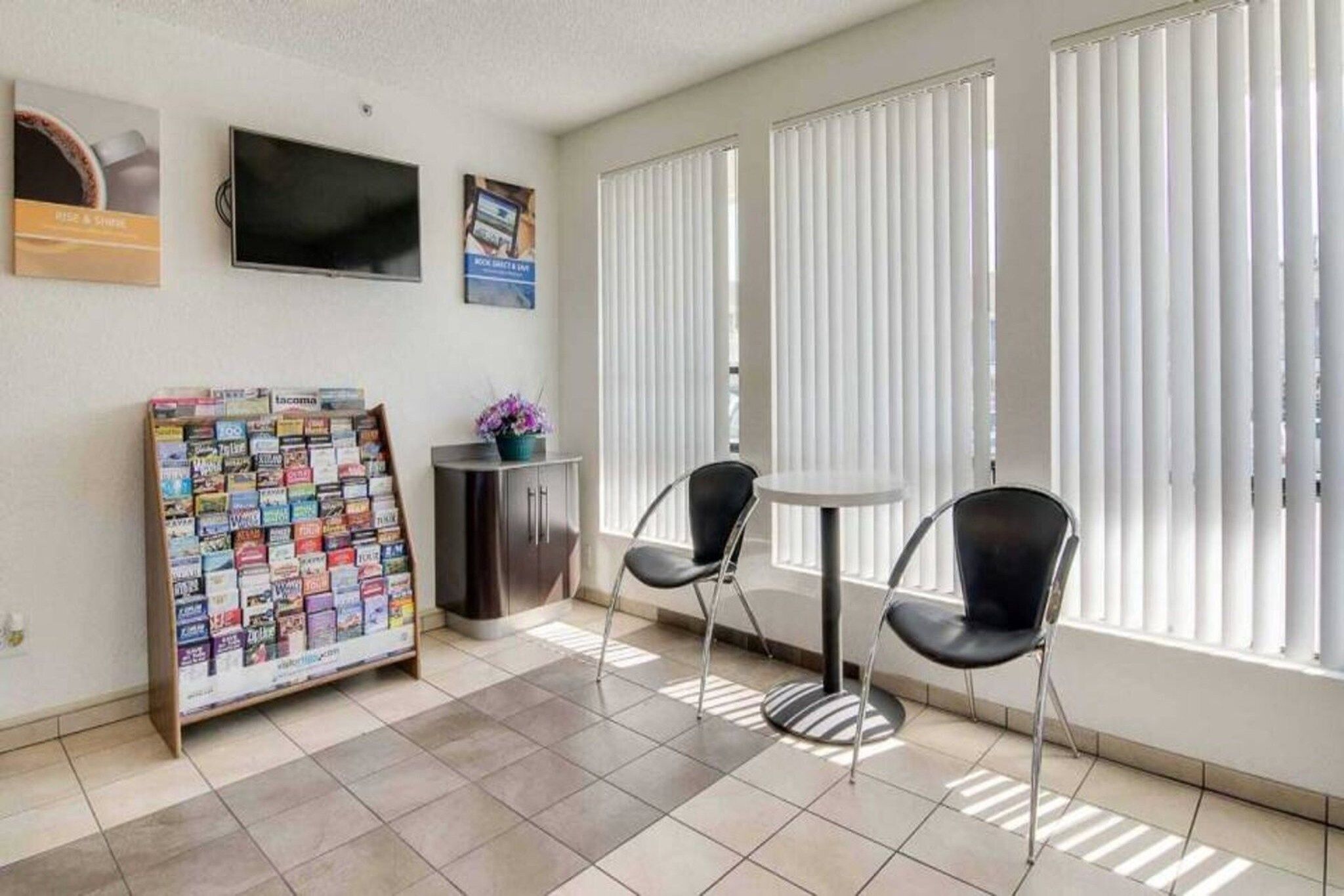 A room with a tiled floor, two black chairs, a small table, a cabinet, a bulletin board, and a window with blinds.