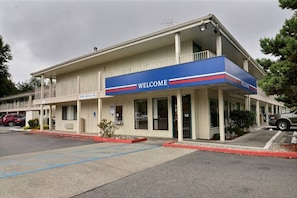 Un motel à deux étages, avec une enseigne « BIENVENUE », un espace de stationnement couvert et une adresse bien visible.