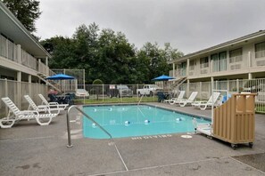 Une piscine entourée d’une clôture et d’arbres, avec des chaises longues et des parasols à proximité.