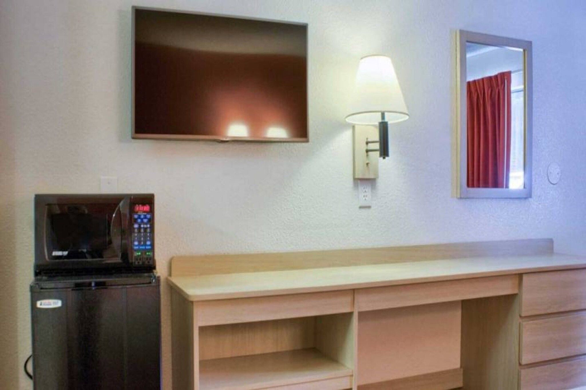A hotel room with a flat-screen TV mounted on the wall, a microwave on a counter, a desk with drawers, a wall-mounted lamp, and a mirror with a red curtain.