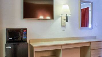 A hotel room with a flat-screen TV mounted on the wall, a microwave on a counter, a desk with drawers, a wall-mounted lamp, and a mirror with a red curtain.