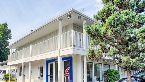 A two-story motel with a balcony on the upper floor, a '6' sign on the front door, and a parking lot in front. - 1 Queen Bed | Micfridge, Non-Smoking (Seattle)