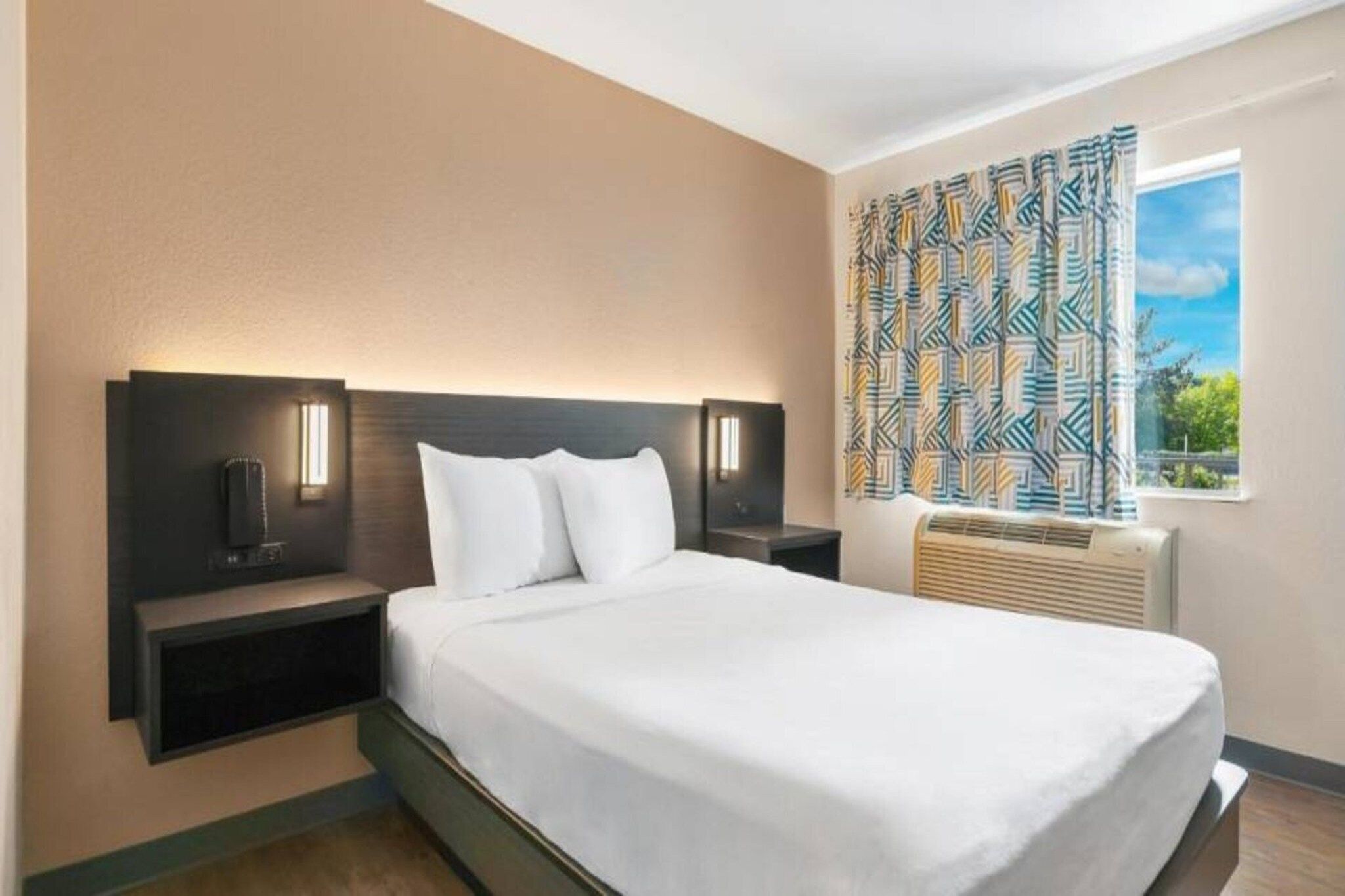 A hotel room with a large bed, two bedside tables with lamps, a window with patterned curtains, and a built-in air conditioning unit.
