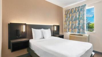 A hotel room with a large bed, two bedside tables with lamps, a window with patterned curtains, and a built-in air conditioning unit.