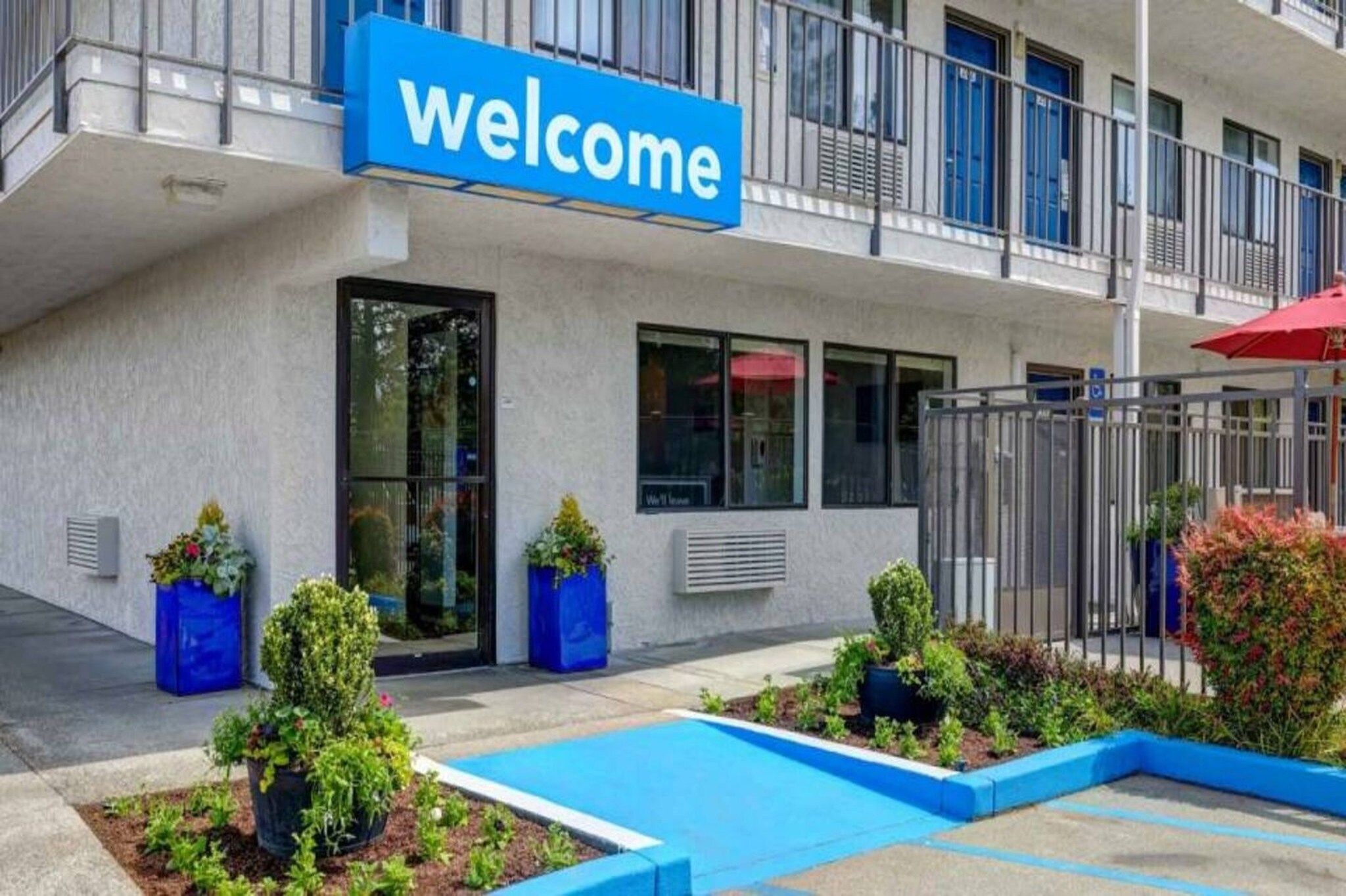 Un bâtiment avec une pancarte indiquant « Bienvenue », une entrée fermée par un portail et un petit espace paysager agrémenté de plantes et d’une piscine.
