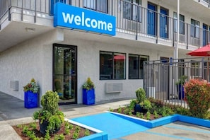 Ein Gebäude mit einem 'Welcome'-Schild, einem Eingangstor und einem kleinen Grünbereich mit Pflanzen und einem Pool.