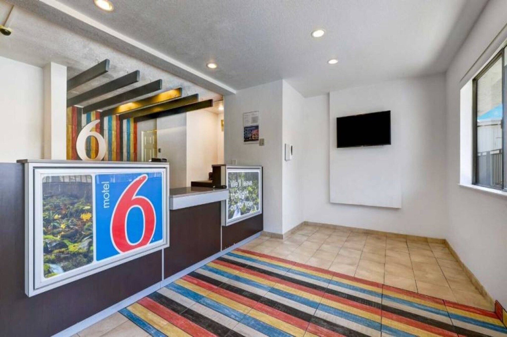 Un hall d’accueil d’hôtel avec un grand panneau portant le chiffre « 6 », un comptoir d’accueil, un téléviseur à écran plat fixé au mur et un tapis rayé aux couleurs vives.