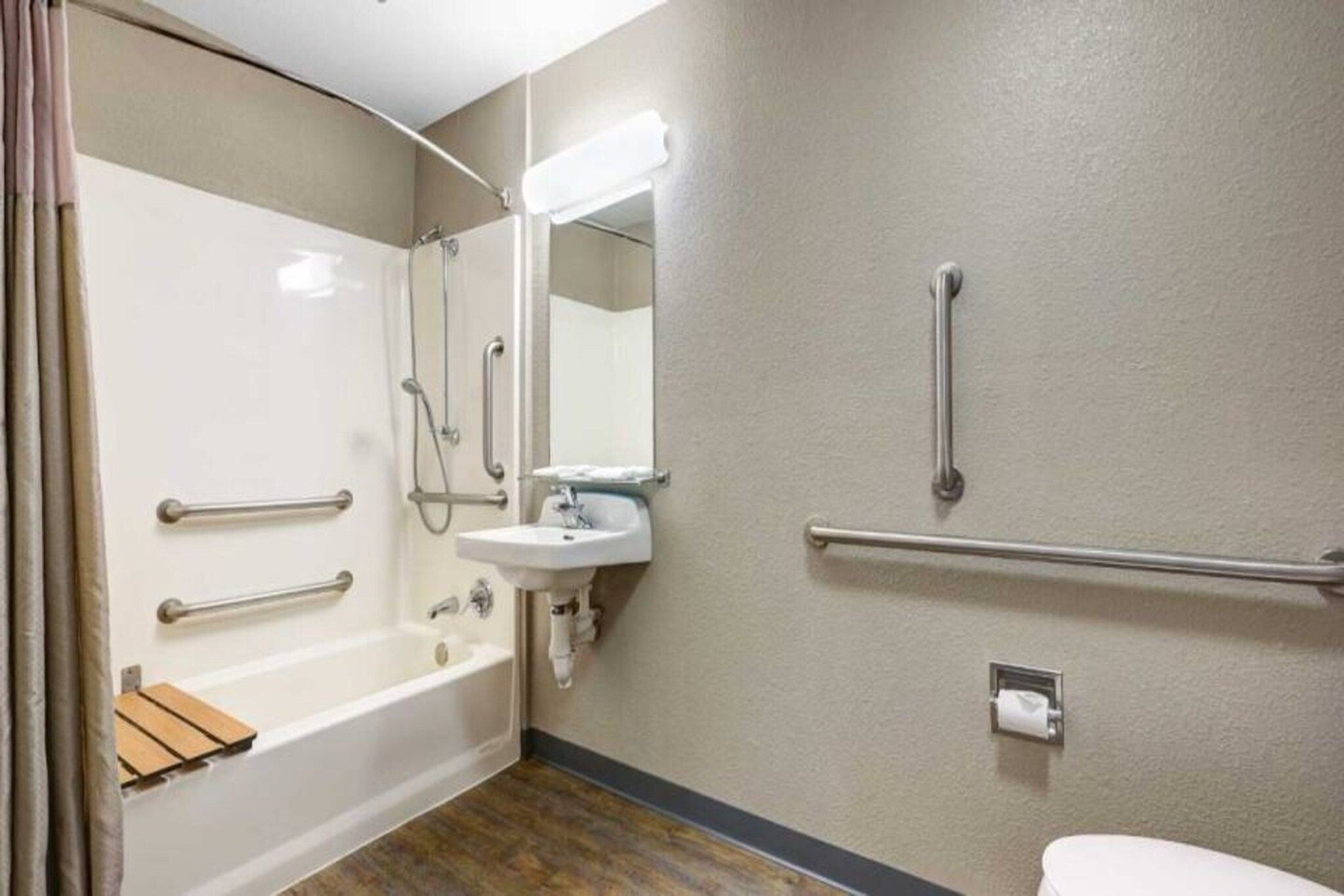 Une salle de bain équipée d’une baignoire, d’un lavabo, d’un miroir et de toilettes. Des barres d’appui sont installées à proximité du lavabo et des toilettes.