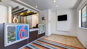 A hotel reception area with a large number '6' sign, a reception desk, a flat-screen TV mounted on the wall, and a colorful striped carpet.