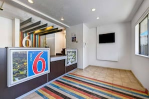 Ein Hotel-Empfangsbereich mit einer Rezeption, einem großen Schild mit der Zahl 6, einem farbenfrohen Streifen Teppich und einem an der Wand befestigten Fernseher.