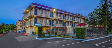 A multi-story hotel with a 'welcome' sign, outdoor seating, and a fenced parking area.