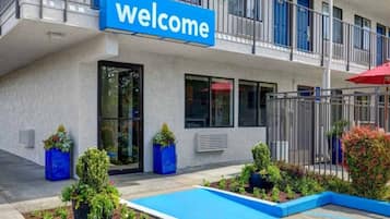 Un bâtiment avec une pancarte indiquant « Bienvenue », une entrée fermée par un portail et un petit espace paysager agrémenté de plantes et d’une piscine.