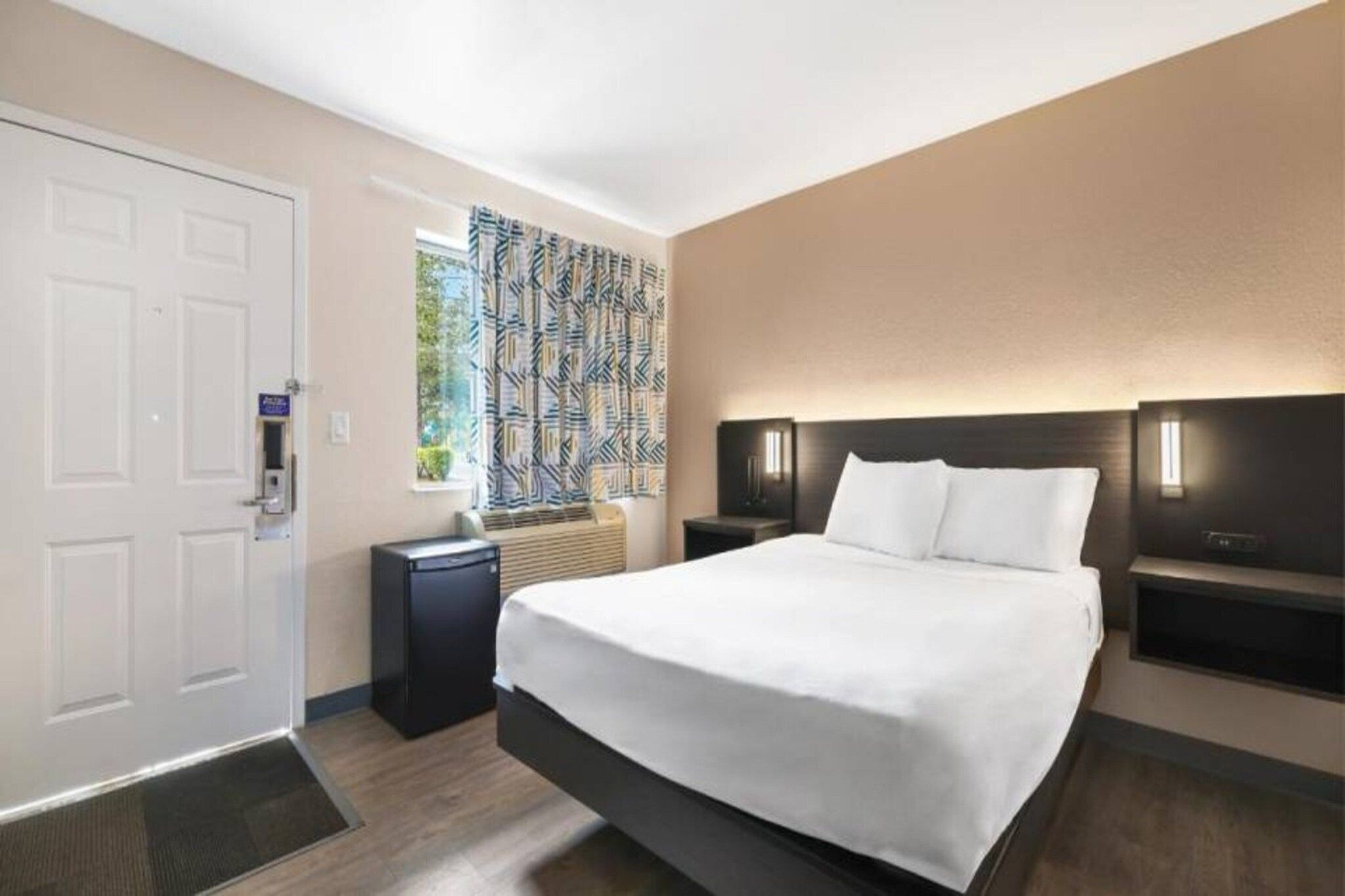 A hotel room with a bed, bedside tables, a heater, a window with patterned curtains, and a door.