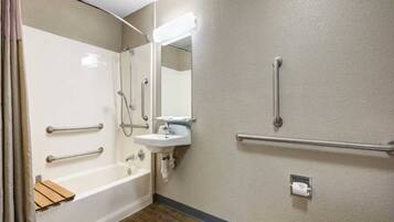 Une salle de bain équipée d’une baignoire, d’un lavabo, d’un miroir et de toilettes. Des barres d’appui sont installées à proximité du lavabo et des toilettes.