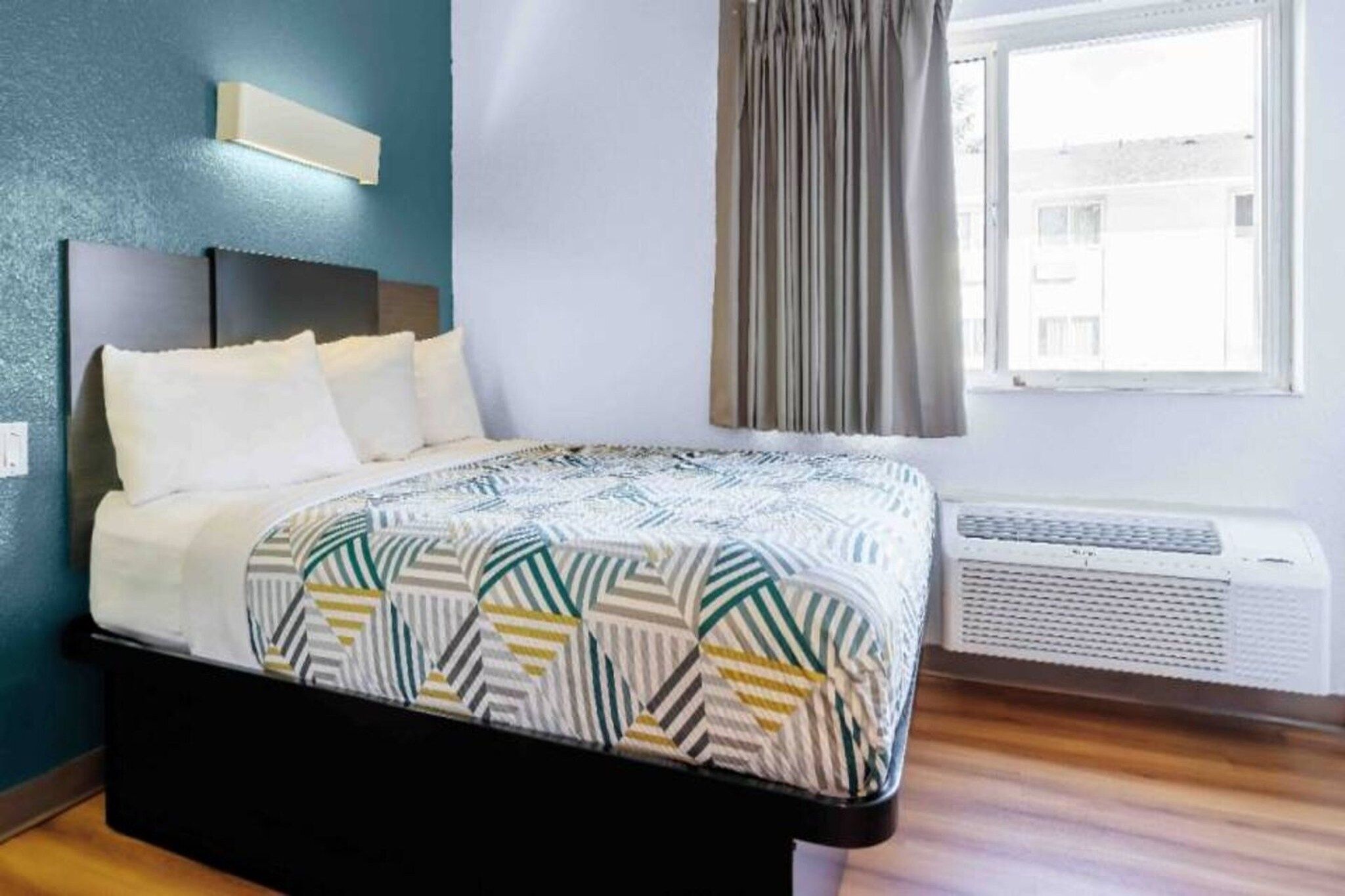 A hotel room with a bed, a headboard, a window with curtains, and an air conditioning unit.