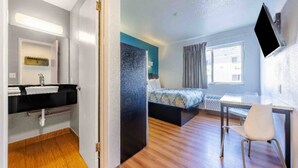 Une chambre d’hôtel avec un plancher en bois, un lit recouvert d’une couvre-lit à motifs, un bureau avec une chaise, un téléviseur à écran plat fixé au mur et une salle de bain avec un lavabo et un miroir.