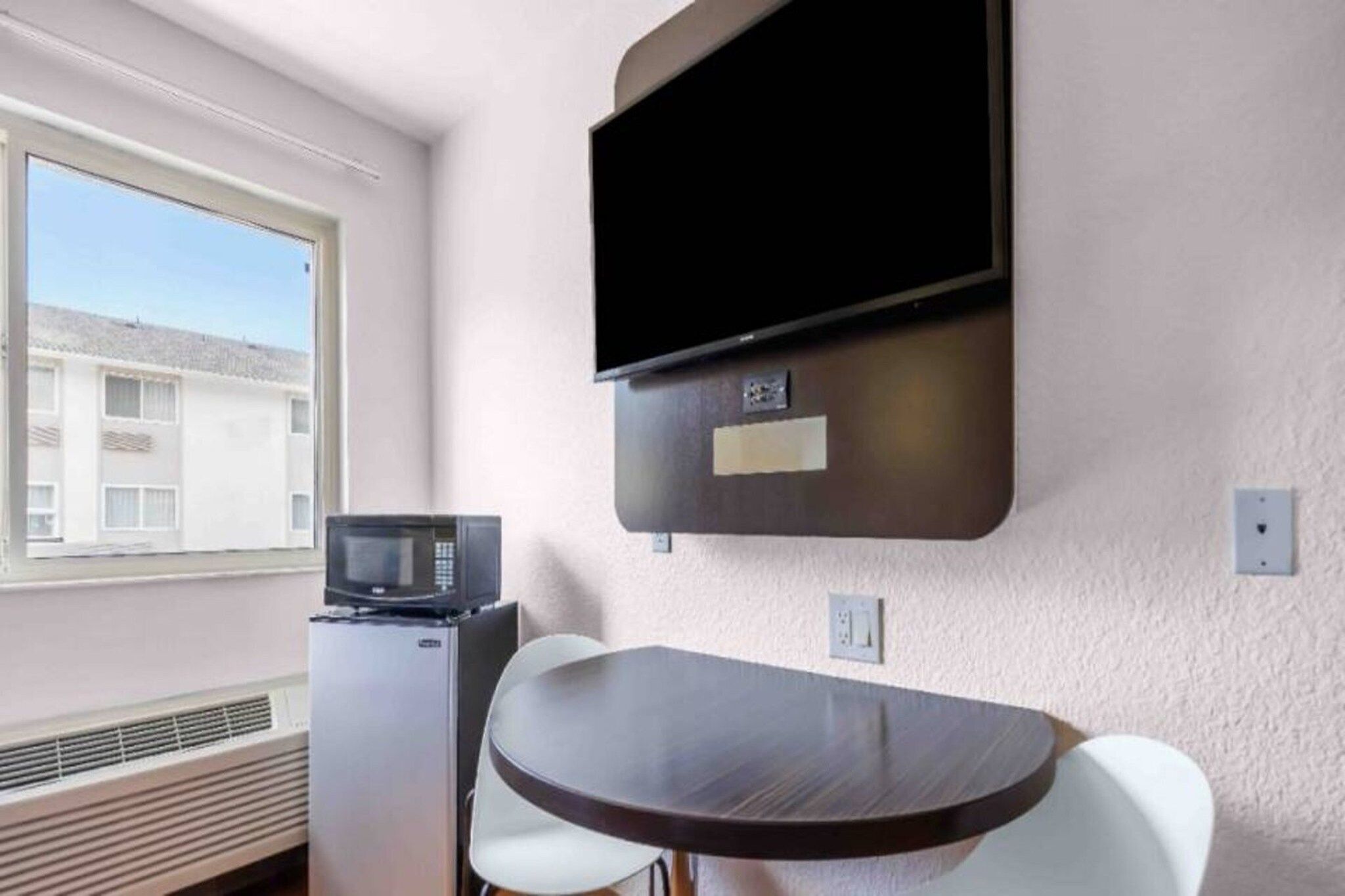 A small room with a flat-screen TV mounted on the wall, a microwave, a small refrigerator, and a round table with two chairs.