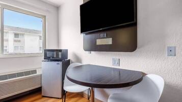 A compact room with a dining table, a microwave, a small refrigerator, a flat-screen TV mounted on the wall, and a window with a view of buildings.