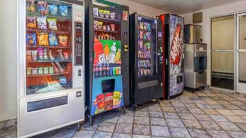 A row of vending machines with snacks and drinks, a refrigerator, and a tiled floor.