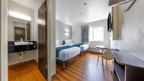 A hotel room with a wooden floor, two beds, a desk, a chair, a TV, and a bathroom with a mirror and sink. - 2 Full Beds | Non-Smoking, Micfridge (Seattle)