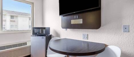 Habitación compacta con mesa de comedor, microondas, pequeño refrigerador, televisor de pantalla plana en la pared y ventana con vista a edificios.