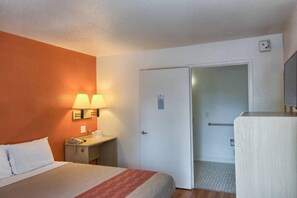 A hotel room with a bed, a bedside table with a lamp, a door, and a small desk. - 1 Queen Bed | Micfridge (Everett)