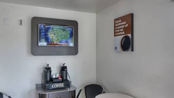 Habitación de hotel con televisor de montaje en pared mostrando un mapa del tiempo, una estación de café y un cartel que ofrece café gratis por la mañana.