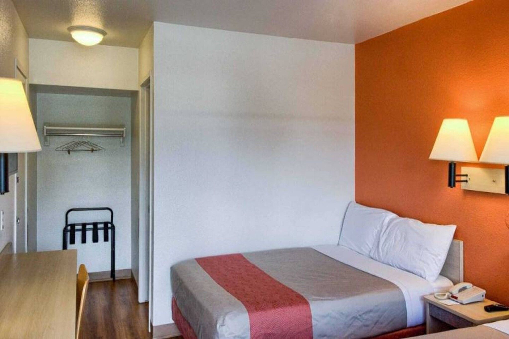 A hotel room with a bed, a desk, and a chair. There is a wall-mounted lamp and a phone on the bedside table.