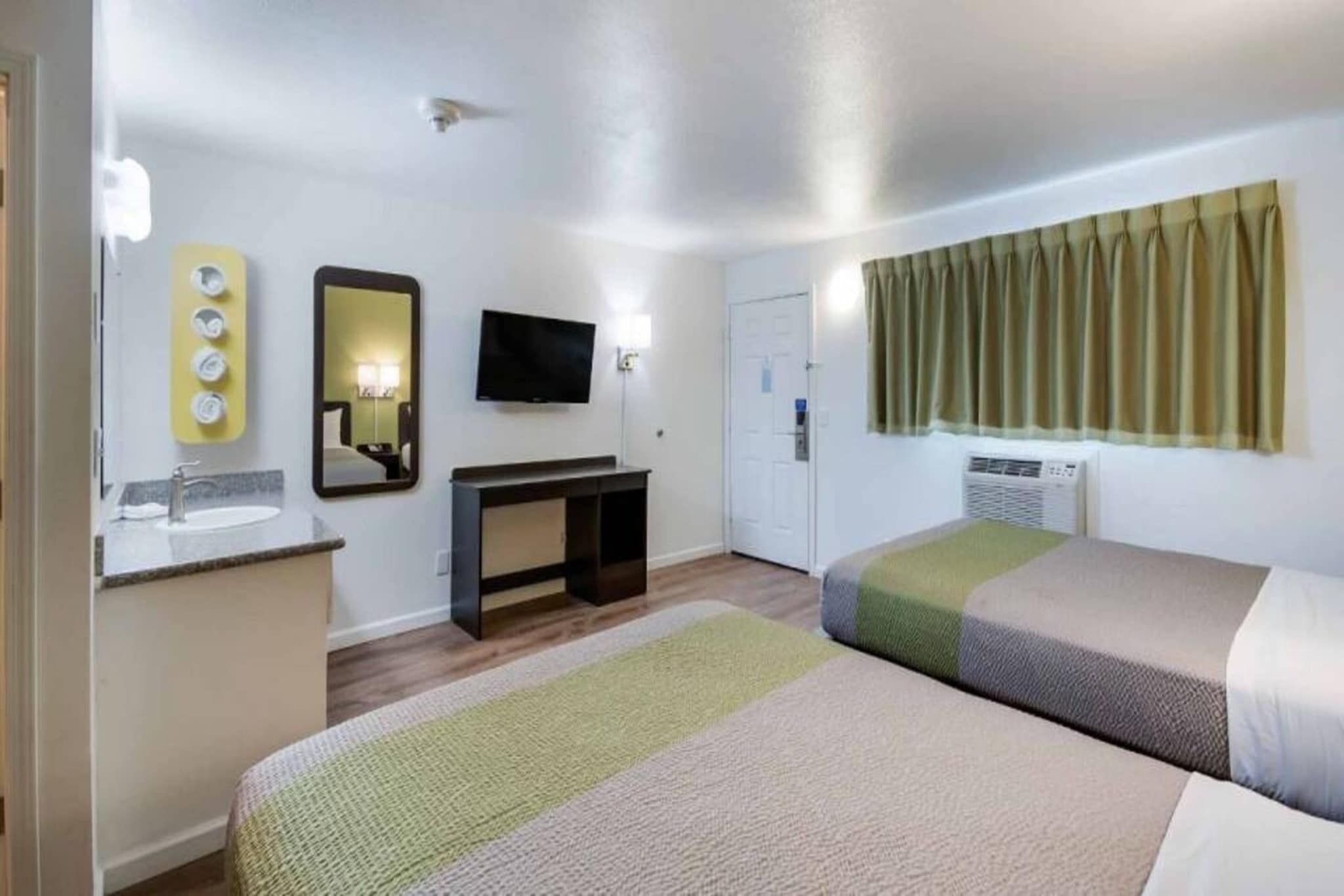 Quarto de hotel com cama, escrivaninha, televisão, espelho, janela com cortinas e banheiro.