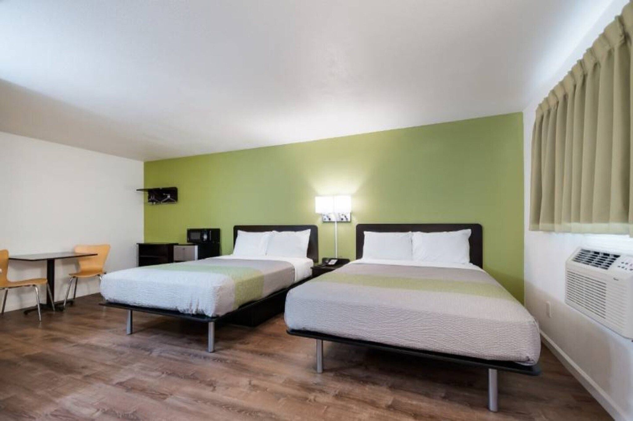 A hotel room with two beds, a desk, a chair, a wall-mounted TV, and an air conditioning unit.