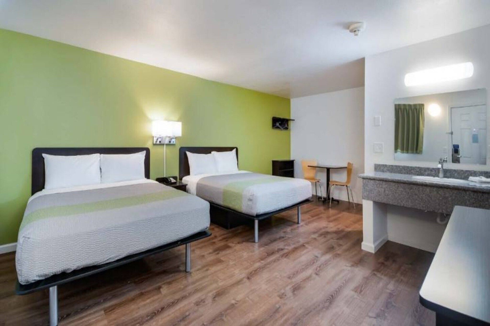 Ein Hotelzimmer mit zwei Betten, einem kleinen Tisch, Stühlen, einem Badezimmer mit Spiegel und einer grünen Wand.