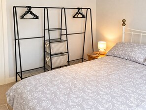 En snyggt bäddad säng med ett blommigt mönstrat överkast, en sänglampa, ett nattduksbord med en bok och en penna samt en hylla i metall med randiga förvaringskorgar.