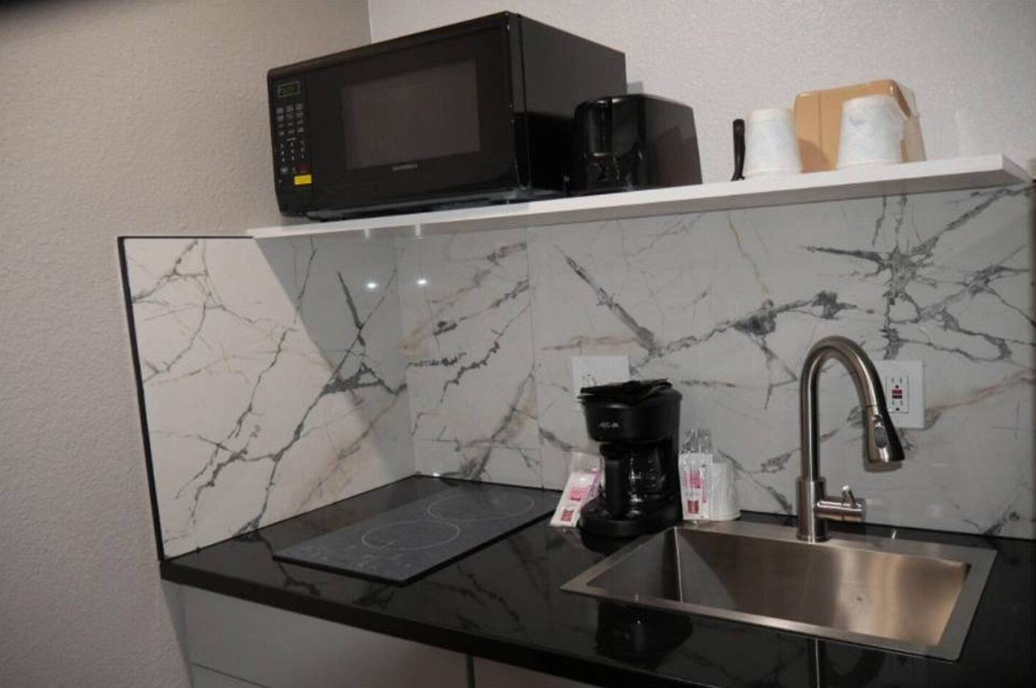 Een keuken met een zwarte magnetron, een koffiezetapparaat en een RVS spoelbak.